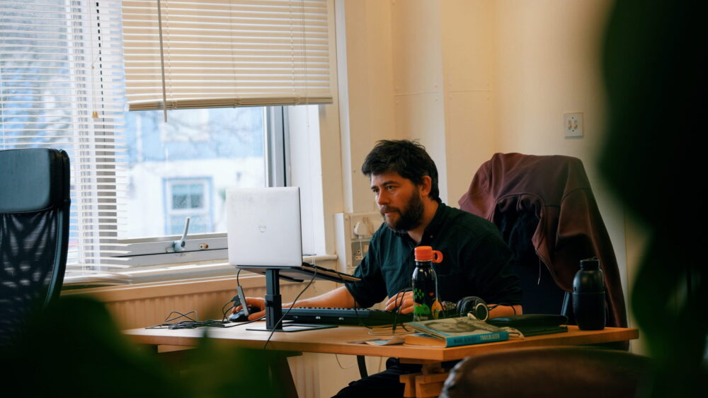 A person sits at a wooden desk in a bright office, working on a laptop connected to an external keyboard and mouse. The desk holds a water bottle, headphones, and a few books. A coat hangs on the back of the desk chair. Sunlight filters through partially closed blinds on a large window beside the workspace.