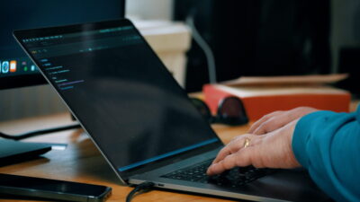 Close-up view of a person typing on a laptop at a wooden desk. The laptop screen displays a dark-themed interface with lines of text. Nearby, there is a smartphone, a monitor, and a pair of sunglasses resting beside a red box.