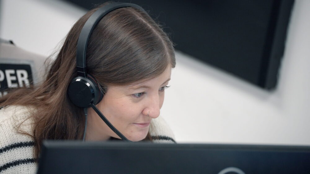 A person wearing over-ear headphones sits at a desk, partially obscured by a computer monitor. Their head is lowered slightly, and long hair falls forward as they work. A soft, neutral-colored wall and part of an office chair are visible in the background.